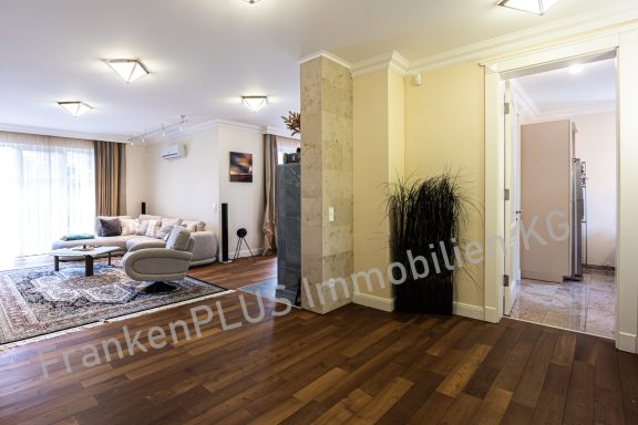 Heller Wohnraum mit Sofa, Teppich und Pflanzen, Zugang zu einem weiteren Raum.