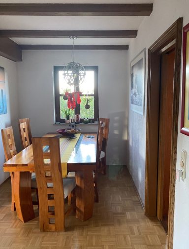 Esszimmer mit Holztisch, Stühlen und einem Fenster, geschmückt mit einem Weihnachtskranz.