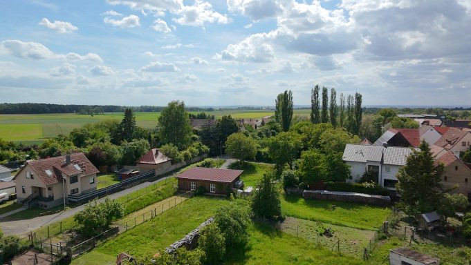 Landschaft mit Wohnhäusern, Bäumen und einem offenen Feld unter bewölbtem Himmel.