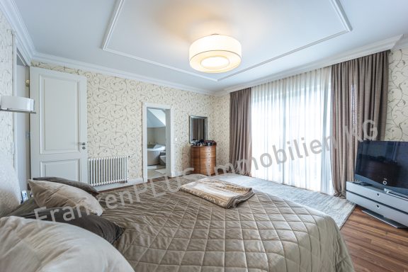 Helles Schlafzimmer mit Doppelbett, Fernseher und großen Fenstern.