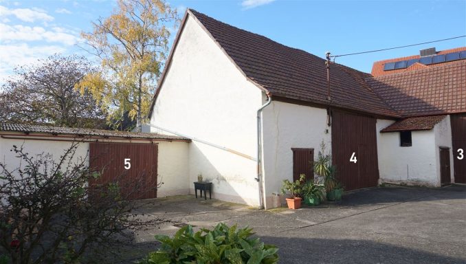 Weißes Gebäude mit braunen Türen und Pflanzen im Vordergrund, blauer Himmel.